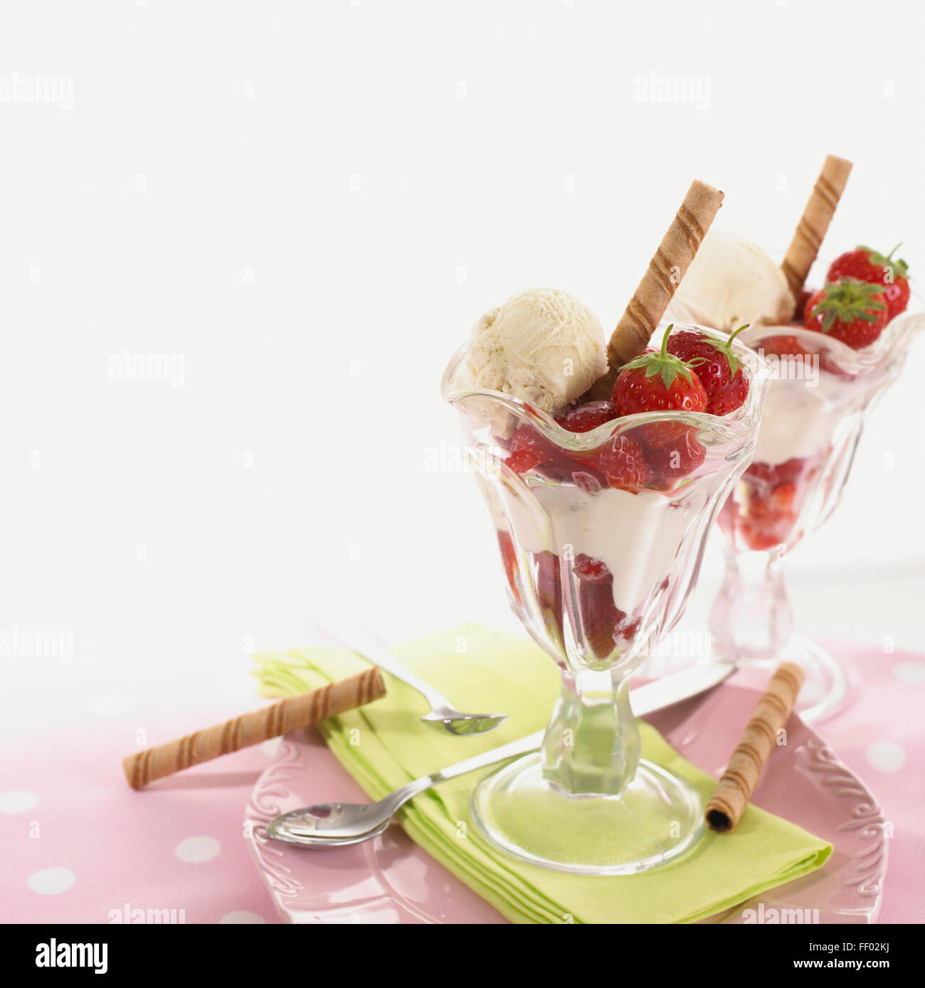 Two servings of vanilla ice cream with fresh strawberries, cream and wafers, served in glasses, with napkin, spoons, on plate, close-up Stock Photo