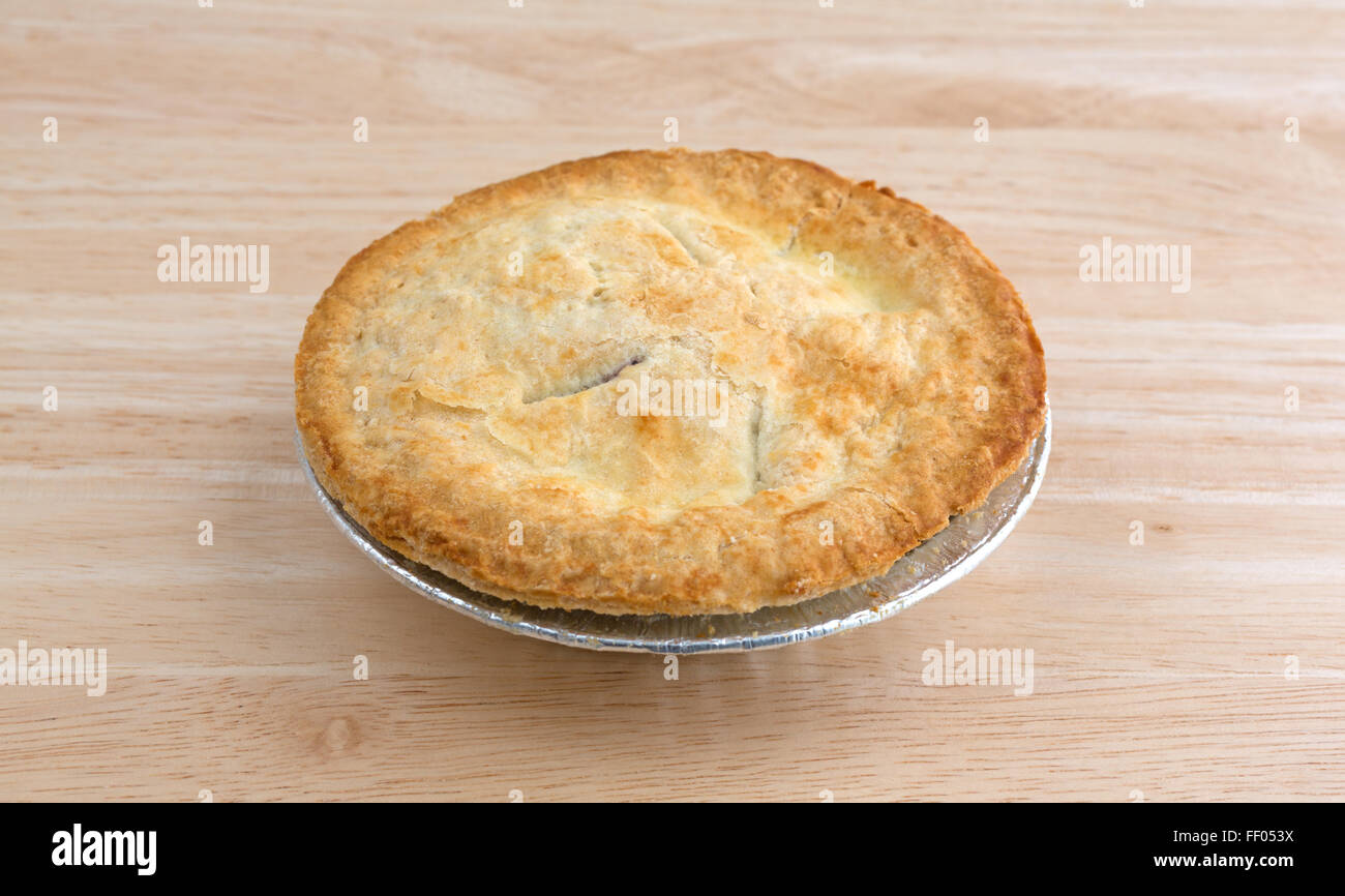 A small personal size fruit filled pie in a tinfoil dish on a wood table top illuminated with natural light. Stock Photo