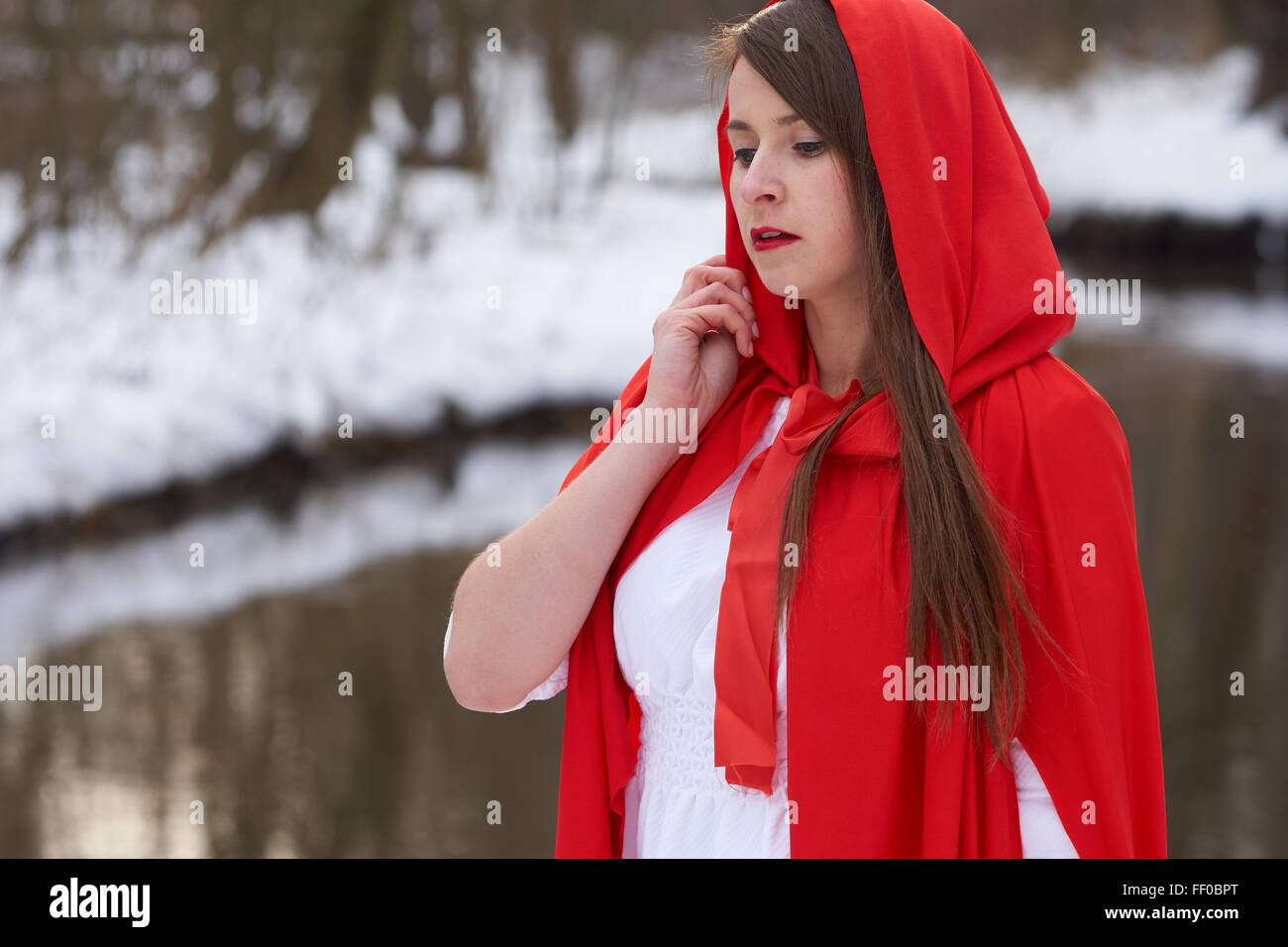One girl in red cape stands near the river in winter Stock Photo