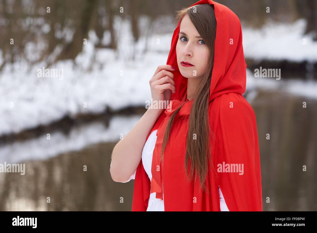 One girl in red cape stands near the river in winter Stock Photo