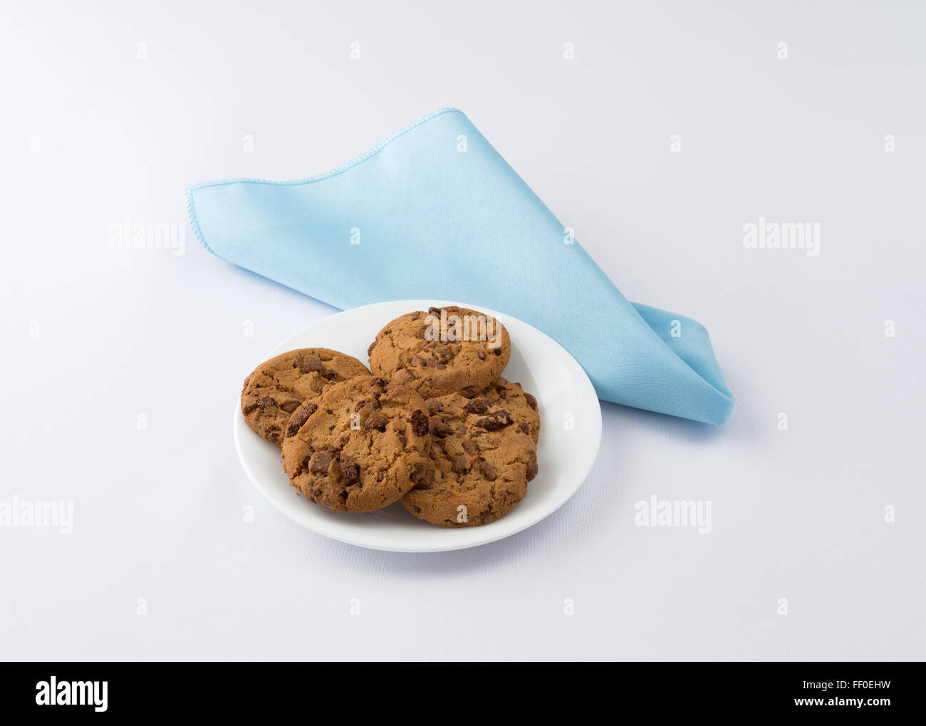 A plate of milk chocolate chip cookies on a white plate with a blue cloth napkin atop an off white tablecloth Stock Photo