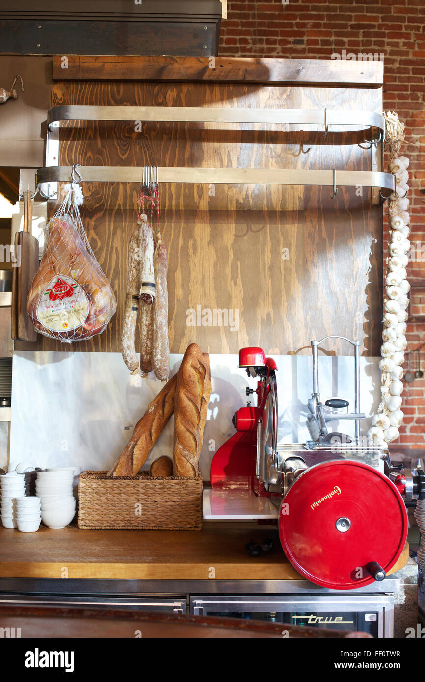 A red Continental meat slicer sitting on a counter with various cured meats, bread and a braid of garlic hanging above. Stock Photo