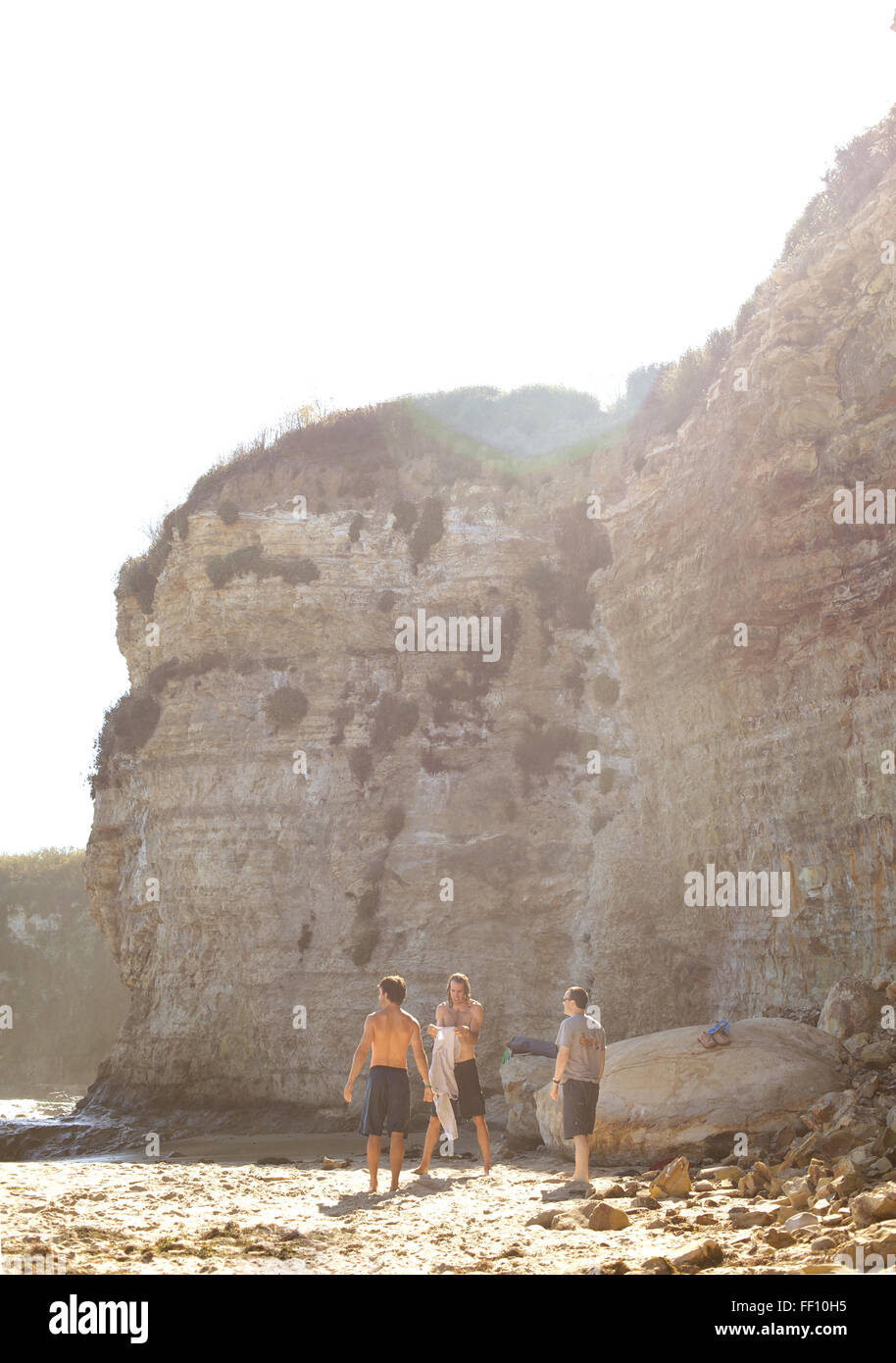Three men standing on a sunny beach with the cliff face in the background, one man puts on his shirt. Stock Photo