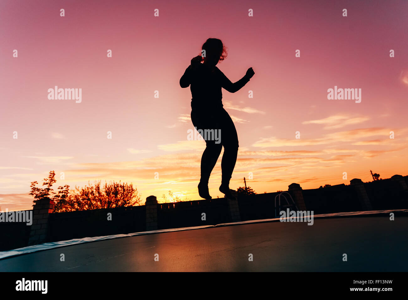 Silhouette Of Beautiful Plus Size Young Woman Girl Jumping On Trampoline On Evening Red, Pink Colorful Sunset Sky Background Stock Photo