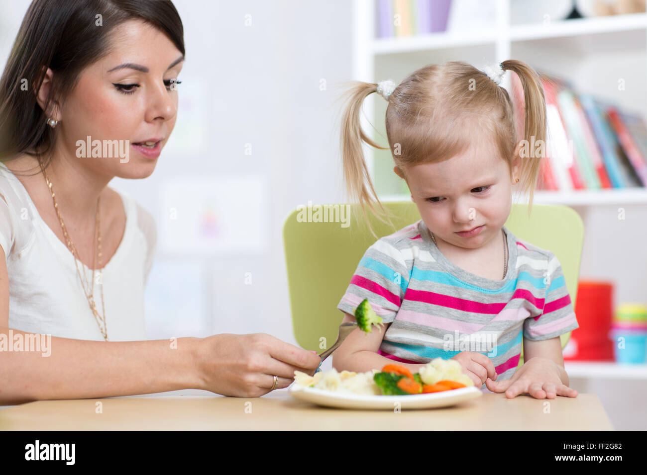 Child girl looks with disgust at healthy vegetables. Mother convinces her daughter to eat food. Stock Photo