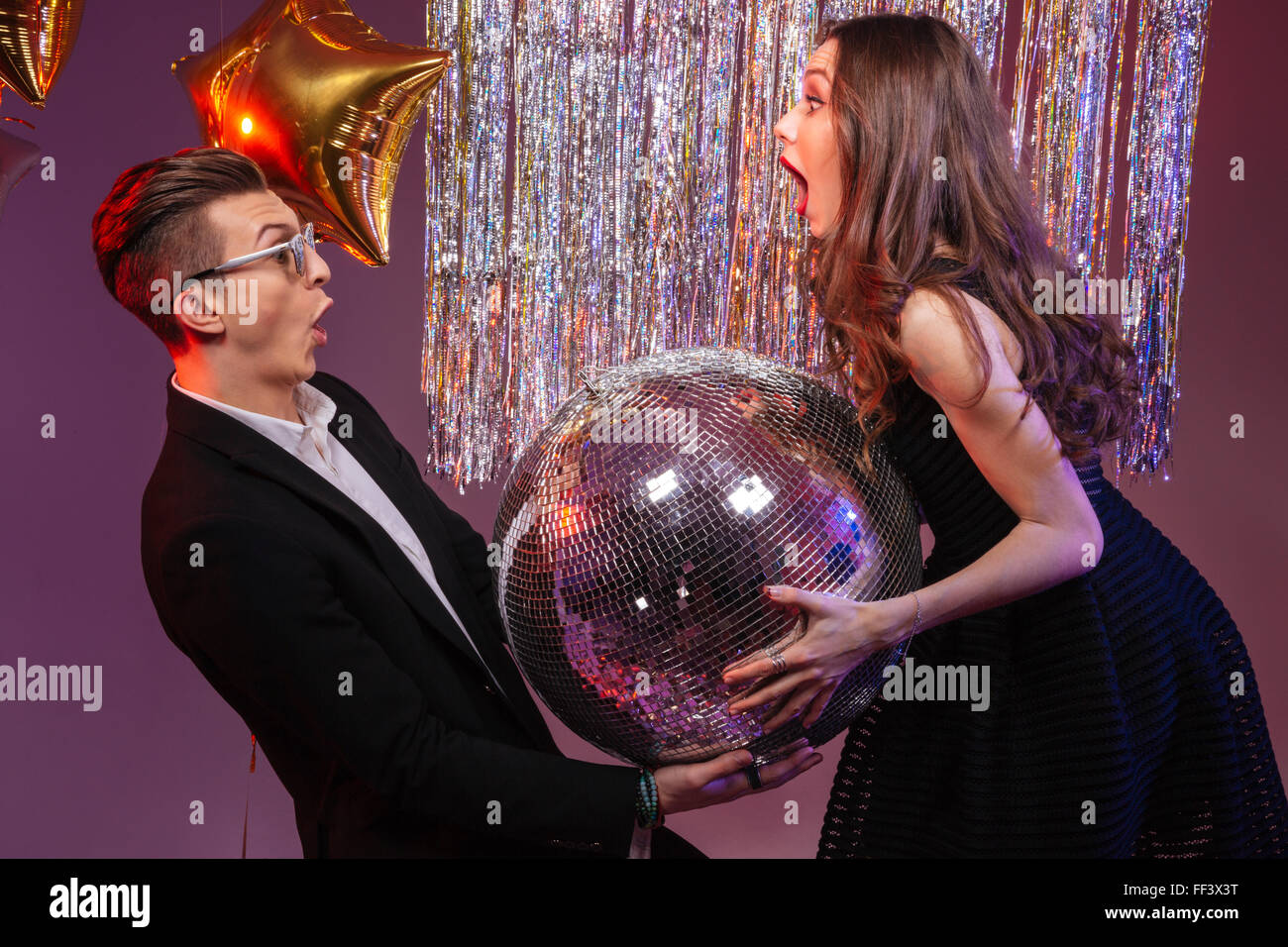 Attractive amazed young couple holding disco ball over purple background Stock Photo