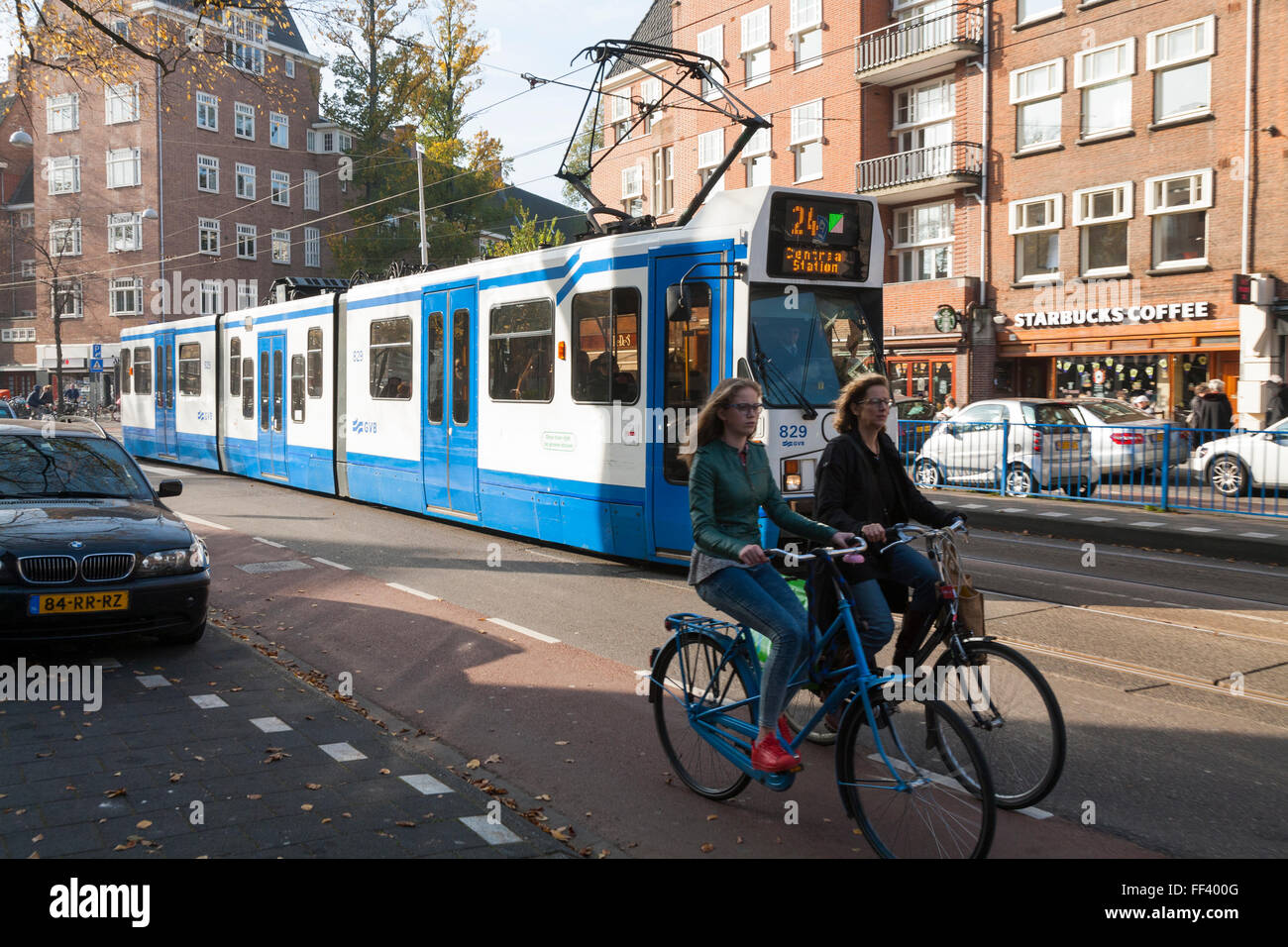 Two cyclists / bike / bikes / cycles and a Line 24 Dutch tram running
