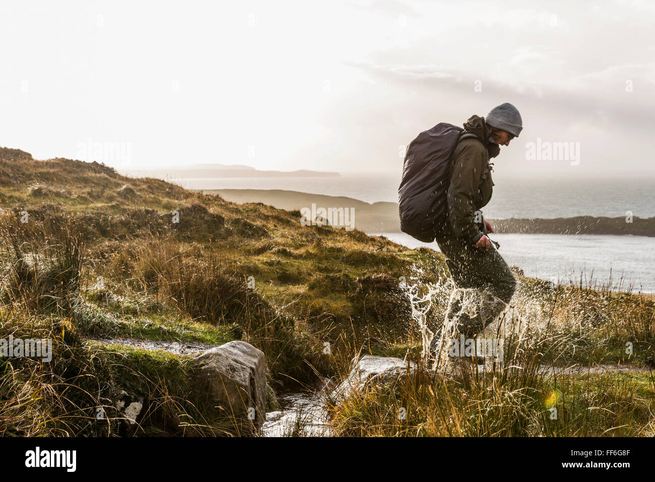 A man with a rucksack and winter clothing leaping across a small stream in an open exposed landscape. Stock Photo