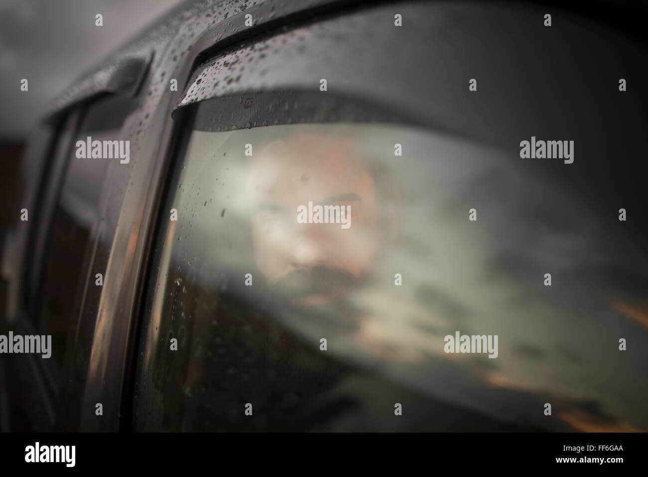 A man sitting in a car looking out. Reflections of the sunset sky on the window. Stock Photo