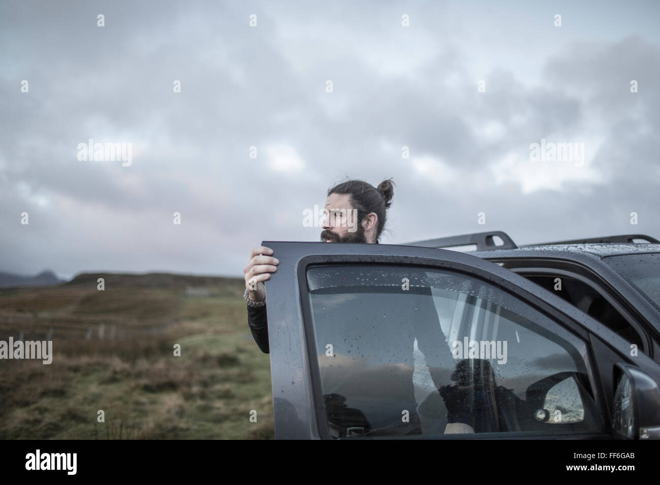 A man standing by an open car door, looking around him, under a cloudy wintery sky. Stock Photo