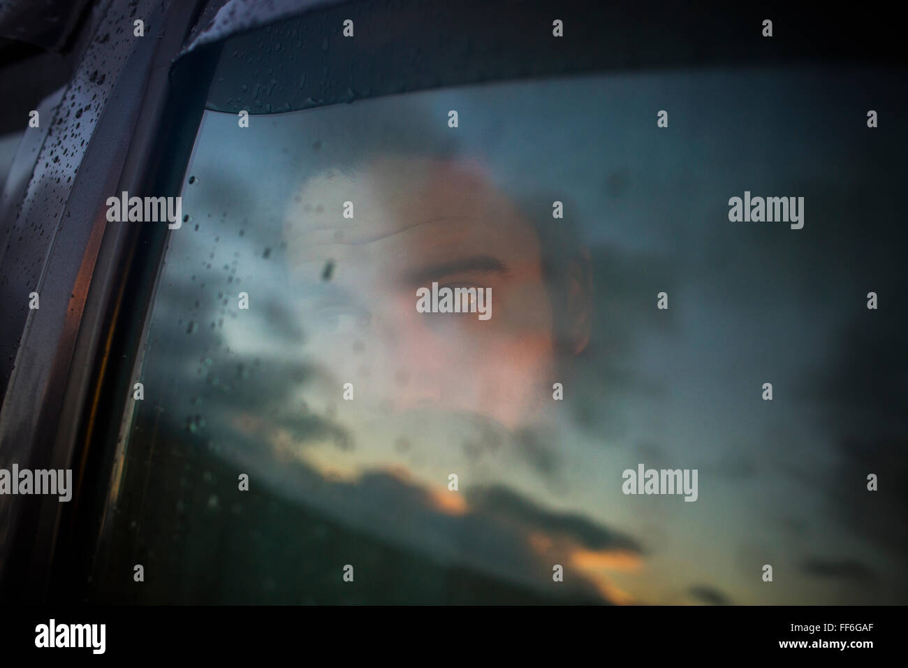 A man sitting in a car looking out. Reflections of the sunset sky on the window. Stock Photo