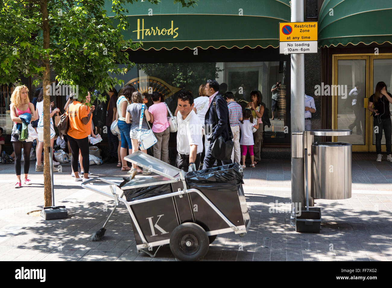 A male street cleaner collecting rubbish and litter outside the world famous and iconic Harrods department store in Knightsbridge, London, United Kingdom.  Groups of tourists are in the background looking at the shop’s window displays being guided around London by Embassy Staff. Stock Photo