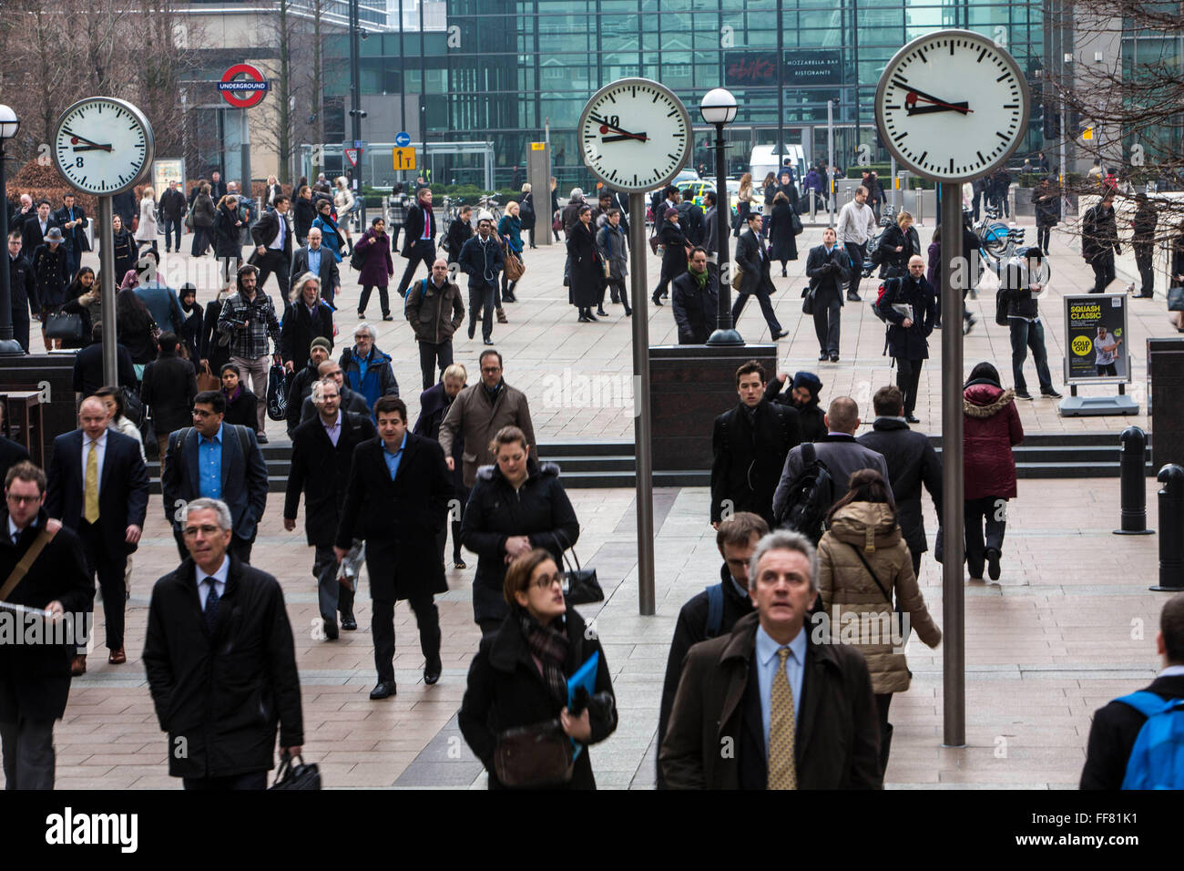 City workers walk under the clocks of Nash Court across Reuters Plaza to commute to work in Canary Wharf financial district London, England, United Kingdom. Stock Photo