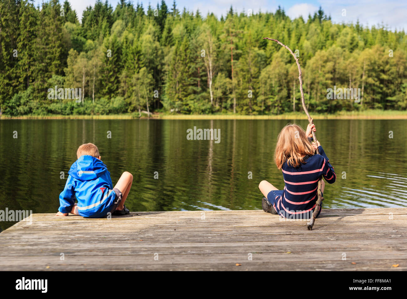 Two children, younger brother and older sister, using a homemade fishing rod fishing from jetty by a lake set in an idyllic Swedish summer forest landscape Stock Photo