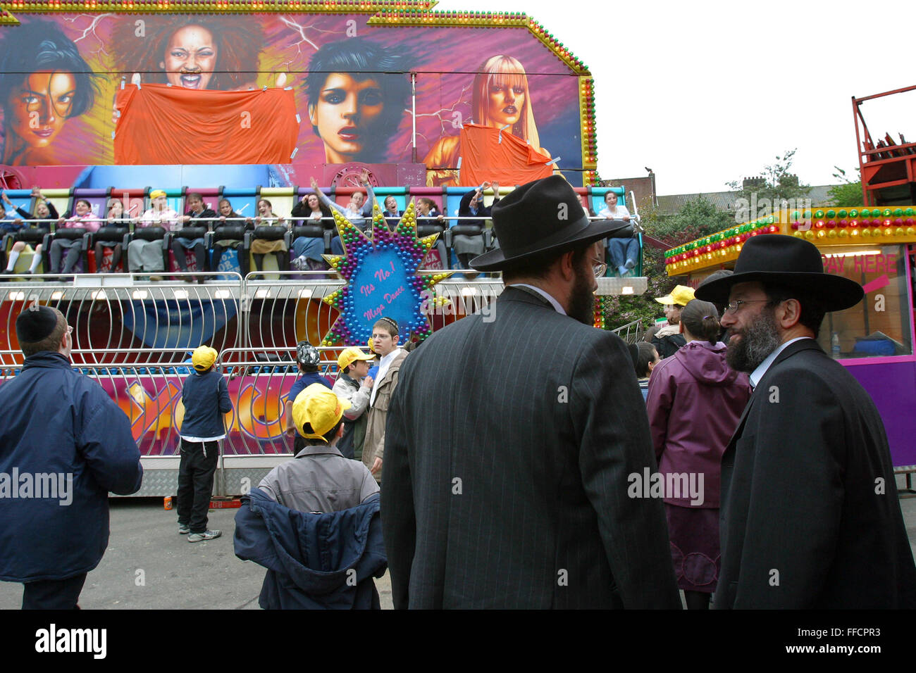 Two men watch their daughters on a fair ground ride in Springfield Park Stamford Hill to celebrate Lag B’Omer. Women and Men are kept separate at all times including children, girls and boys take it in turns to go on the rides. Lag B’Omer is the holiday celebrating the thirty-third day of the (counting of the) Omer. Jews celebrate it as the day when the plague that killed 24,000 people ended in the holy land (according to the Babylonian Talmud). Other sources say the plague was actually the Roman occupation and the 24,000 people died in the second Jewish – Roman war  (Bar Kokhba revolt of the Stock Photo