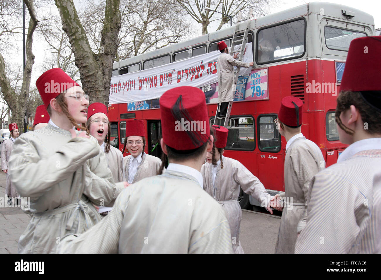 A group of Orthodox Jewish boys from the Viznitz Yeshiva (school) dressed in fancy dress sing and dance in the street. Other members of the group decorate the double-decker bus with banners that they will travel on for the Jewish festival of Purim. They will visit several local wealthy businessmen collecting money for their school. Stock Photo