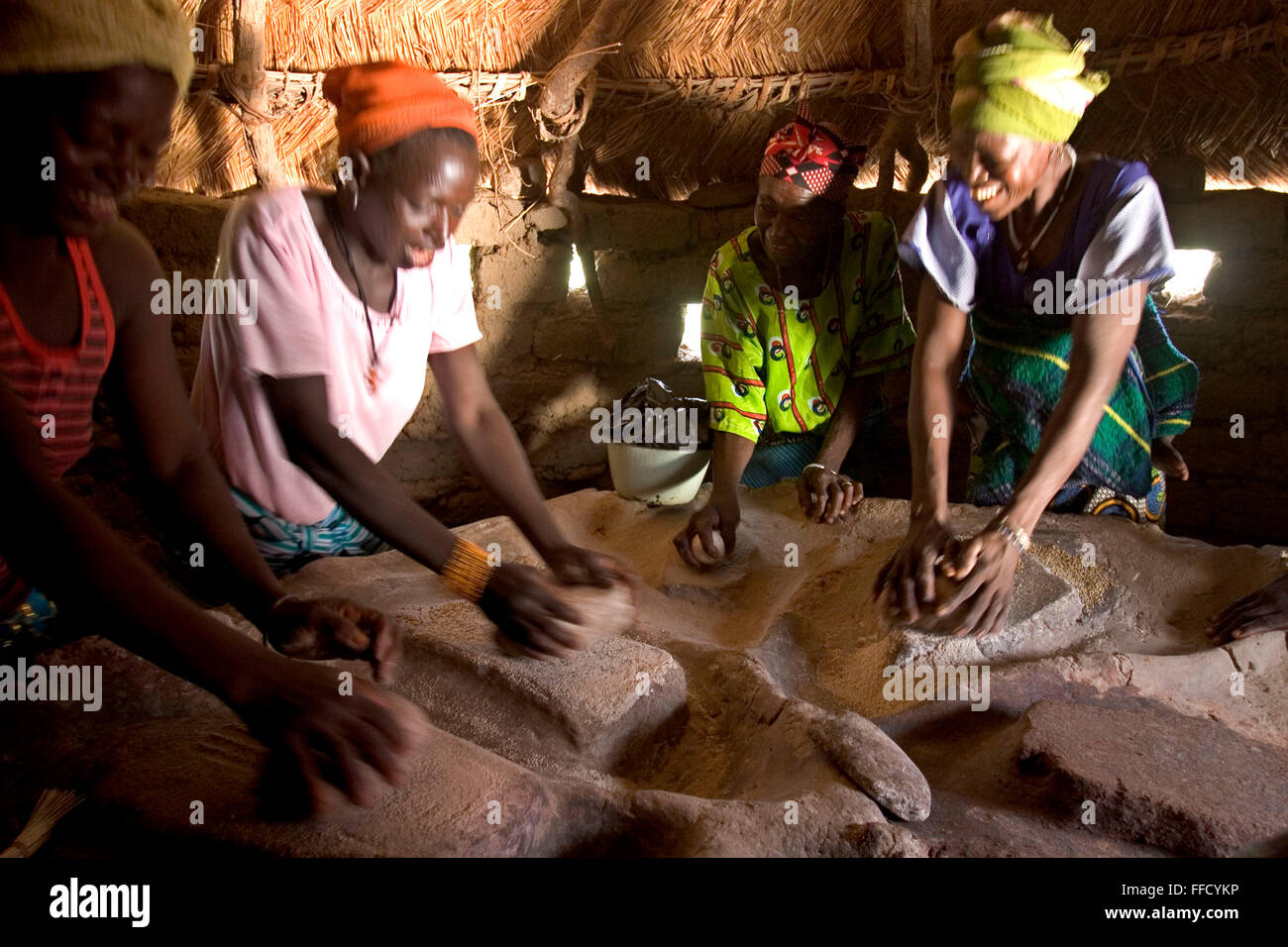 A group of women in Fada N’Gourma Burkina Faso grind organic millet on a traditional table carved from stone to make flour. The table has been in the village for years and has well molded troughs to collect the flour. Stock Photo