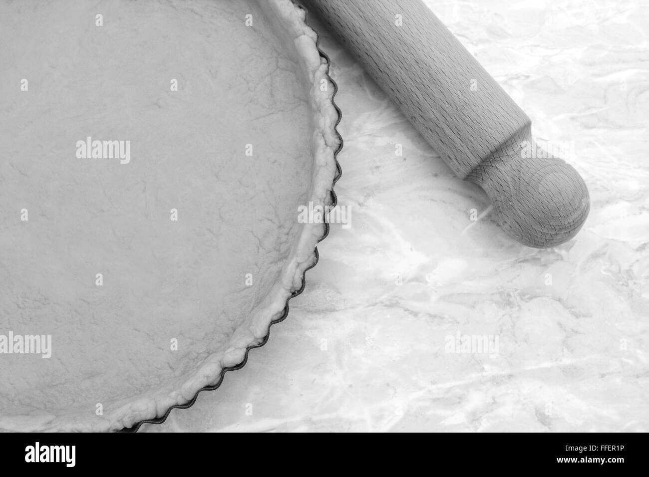 Close-up of circular baking tin lined with pastry, wooden rolling pin on worktop beside - monochrome processing Stock Photo