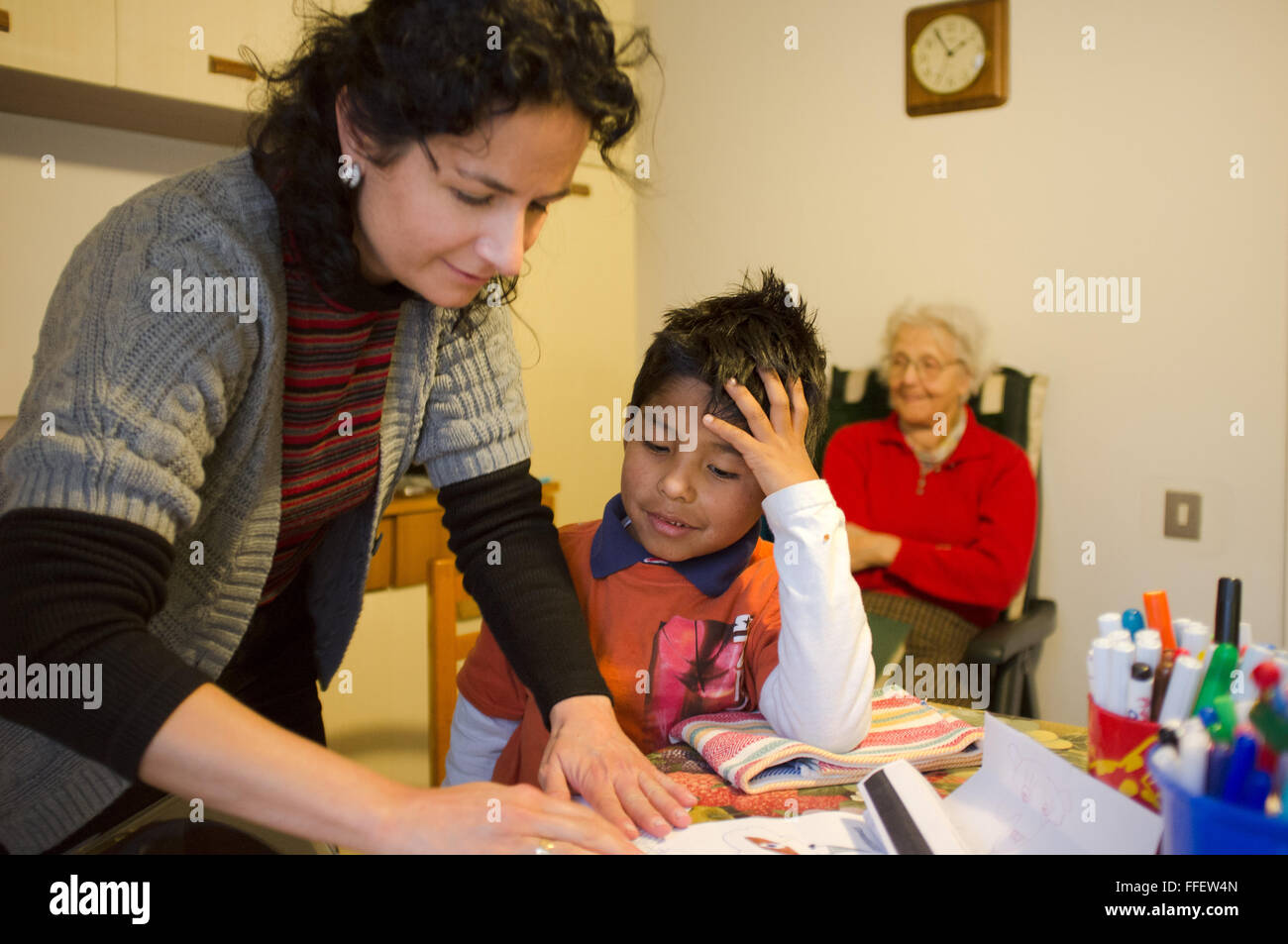 Mature caucasian woman helps her adopted latin boy with his homework while great grandmother watches smiling on a close chair Stock Photo