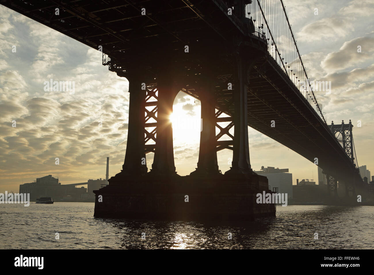 The sun rises under the Manhattan Bridge spanning the East River joining Manhattan to DUMBO, Brooklyn. A power station chimney c Stock Photo