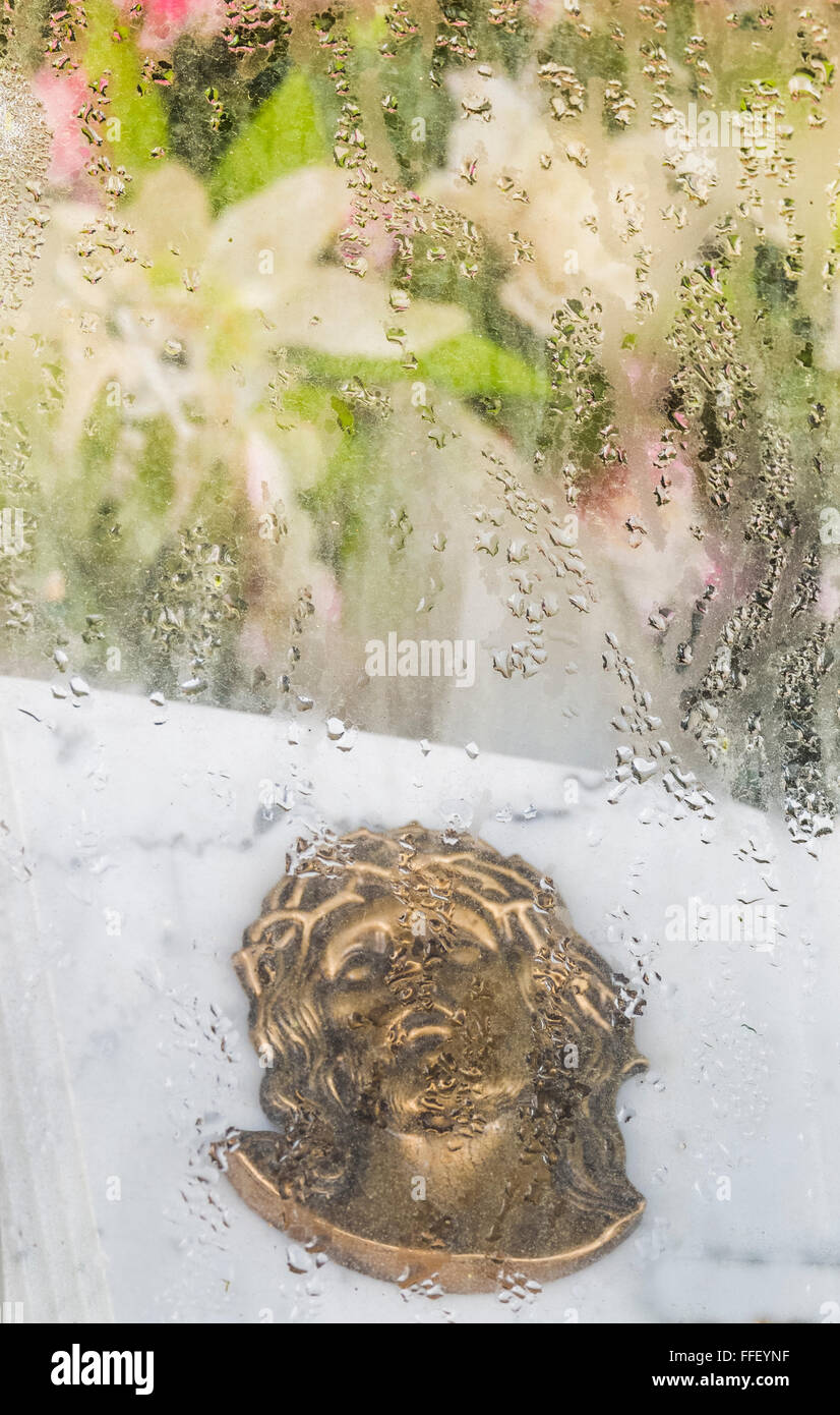 bronze medallion showing jesus christ wearing a crown of thorns behind a moist glass surface light flowers in the background Stock Photo