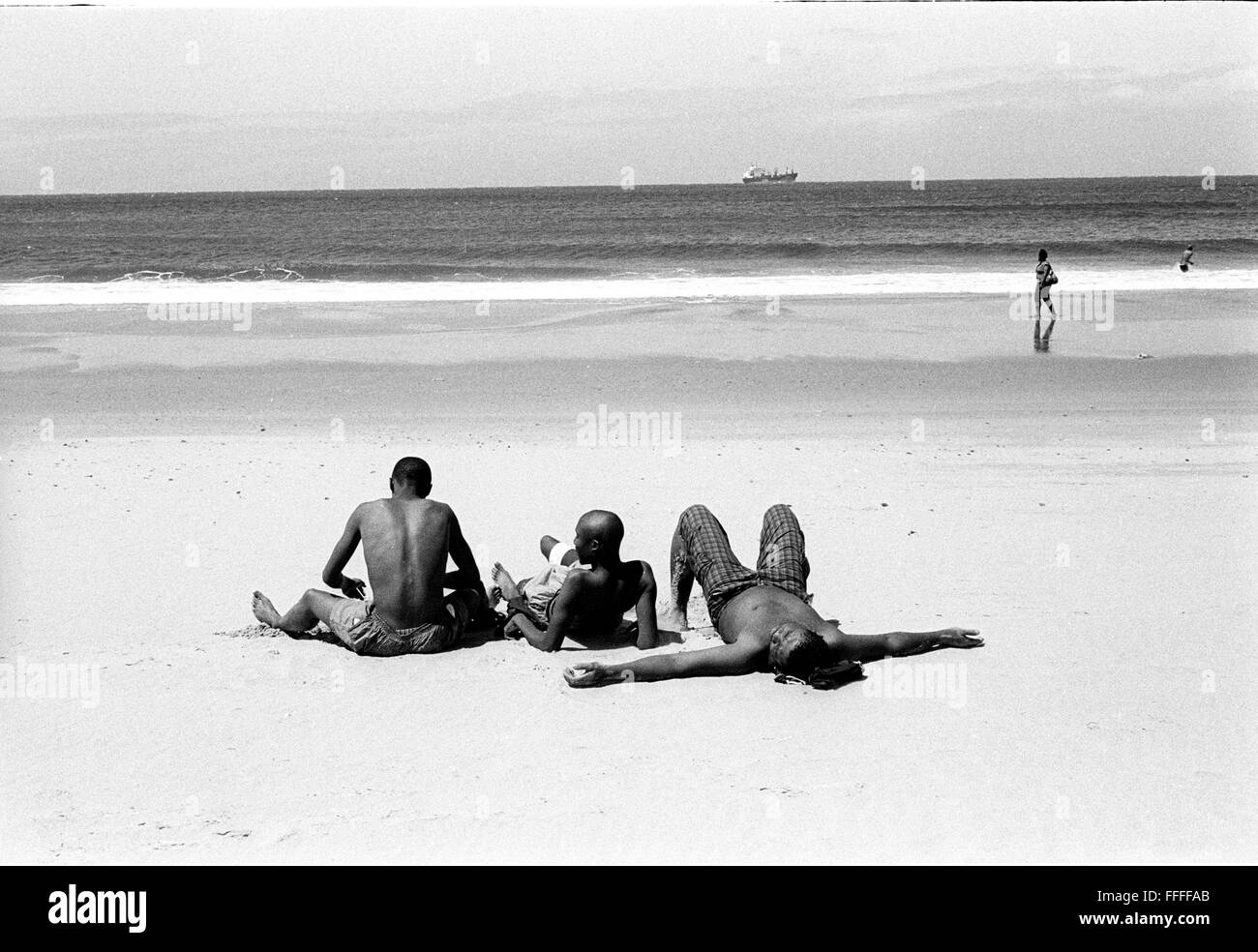 Jan 4, 2016 - South Beach, KwaZulu-Natal, South Africa - Three young men sun bathing with a ship and a man walking in the background. South Beach is a part of the City of Durban's longest uninterrupted stretch of beach sand. The City of Durban is on the eastern seaboard of South Africa and the people here are washed with the warm waters of the Indian Ocean. To the north of this stretch of sand are beaches with cafe society hang outs. To the south there is a pier with the upmarket Moyo's Restaurant at it's end and the uShaka Marine World complex and the private surf and sea clubs of the Vetches Stock Photo