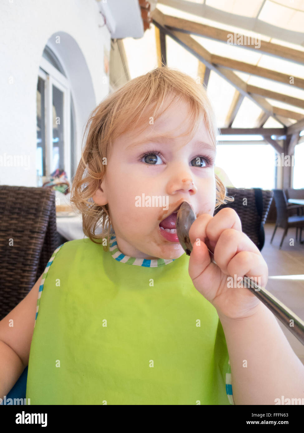face of blonde baby two years old green bib and silver metal spoon in her mouth and smeared with chocolate in restaurant Stock Photo