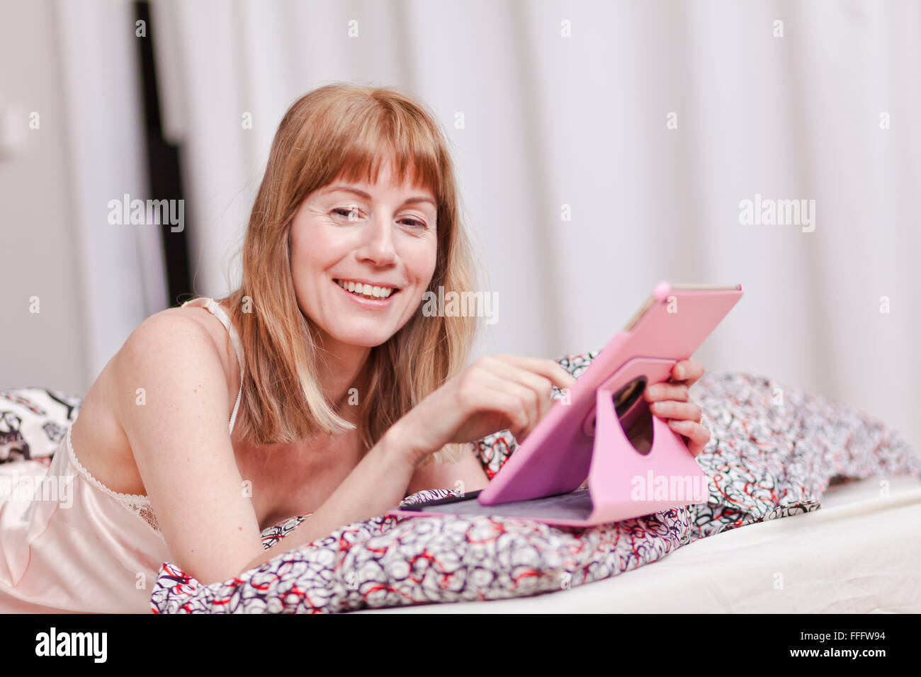 Mid age blond, a caucasian woman on a bed using a pink tablet. Stock Photo