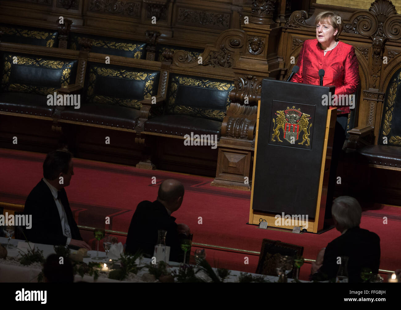 Hamburg, Germany. 12th Feb, 2016. German Chancellor Angela Merkel (back) delivers a speech at the Matthiae dinner, with Britain's Prime Minister David Cameron (L-R), Hamburg's Governing Mayor Olaf Scholz and Michael Otto, former CEO of Otto Group, pictured in the foreground, at the city hall of Hamburg, Germany, 12 February 2016. Britain's Prime Minister David Cameron and German Chancellor Angela Merkel are guests of honour at the oldest feast in the world. Since 1356, the leaders of the Hansa city invite distinguished guests to the Matthiae dinner. Photo: LUKAS SCHULZE/dpa/Alamy Live News Stock Photo