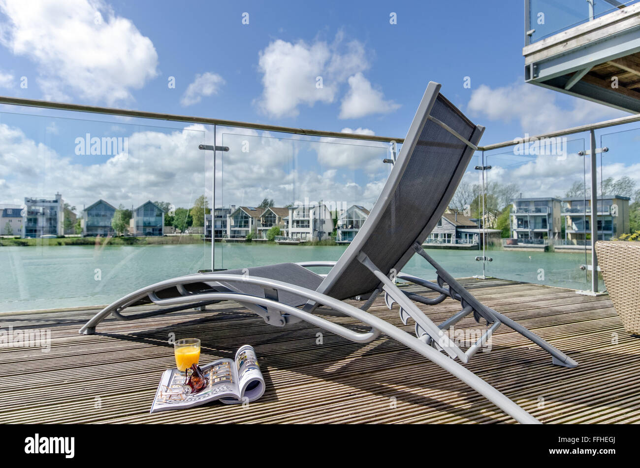 A sun lounger on a decking, waterside veranda overlooking a lake & waterfront homes, South Cerney, Cotswolds, UK Stock Photo