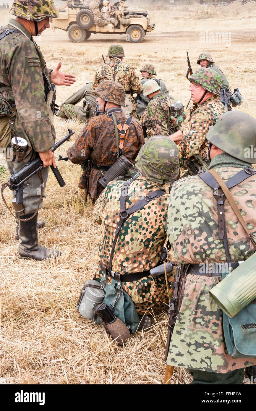 Second world war re-enactment. Group of German Waffen SS soldiers, in dot pattern camouflage uniforms, sitting and waiting in yellow grass. Stock Photo