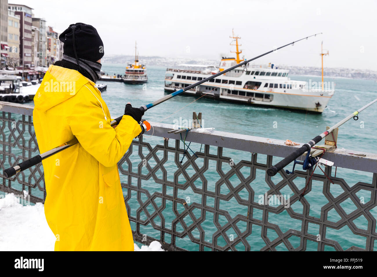 Unidentified local fishermen wearing yellow raincoat fishing on the Galata Bridge on a snowy day in Istanbul, Turkey. Stock Photo