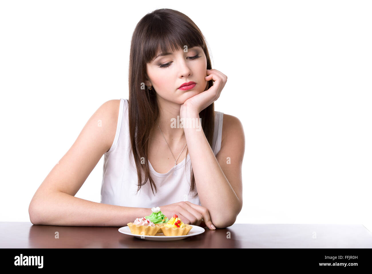 Young dieting woman sitting in front of plate with delicious cream tart cakes in dough baskets, looking at sweet dessert Stock Photo