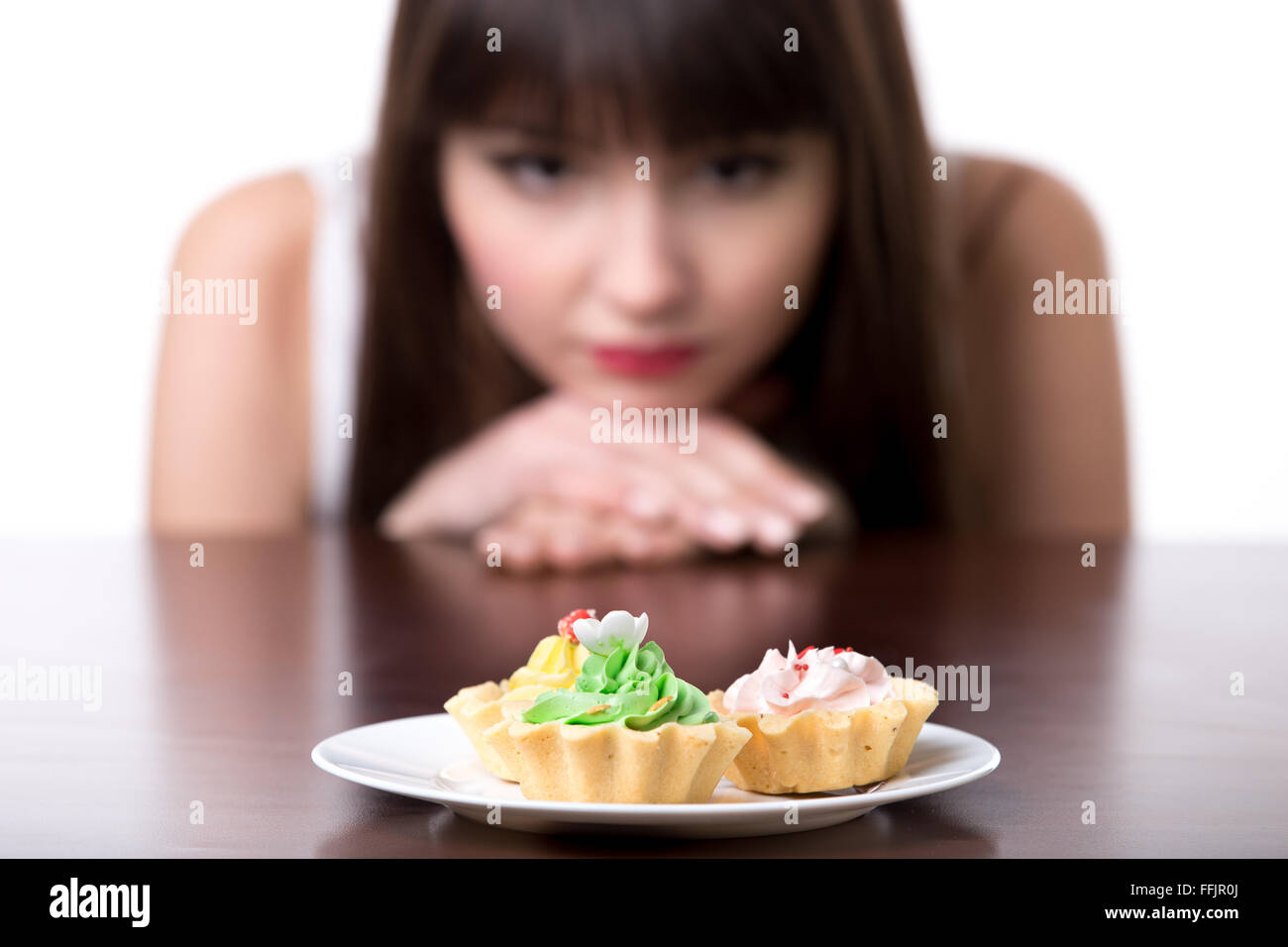 Young dieting woman sitting in front of plate with delicious cream tart cakes, looking at forbidden food with unhappy and hungry Stock Photo