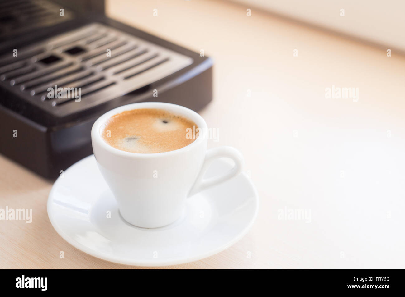 Brewing classic espresso with coffee machine. Coffee with brown froth in small white cup on the kitchen table near window Stock Photo
