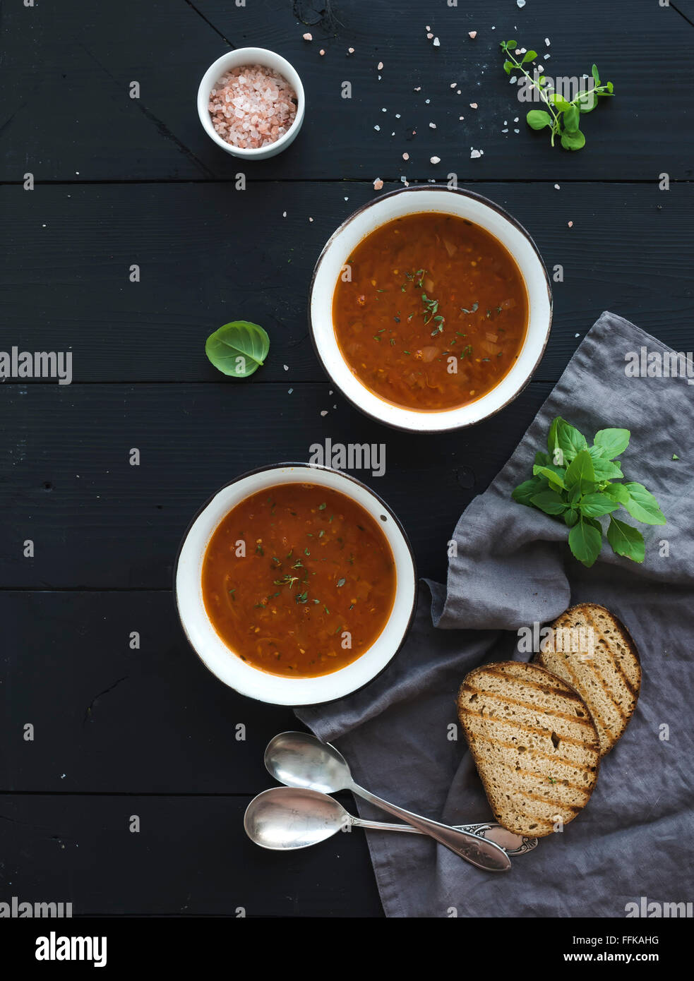 Roasted tomato soup with fresh basil, spices and bread in rustic metal bowls over black background, top view Stock Photo