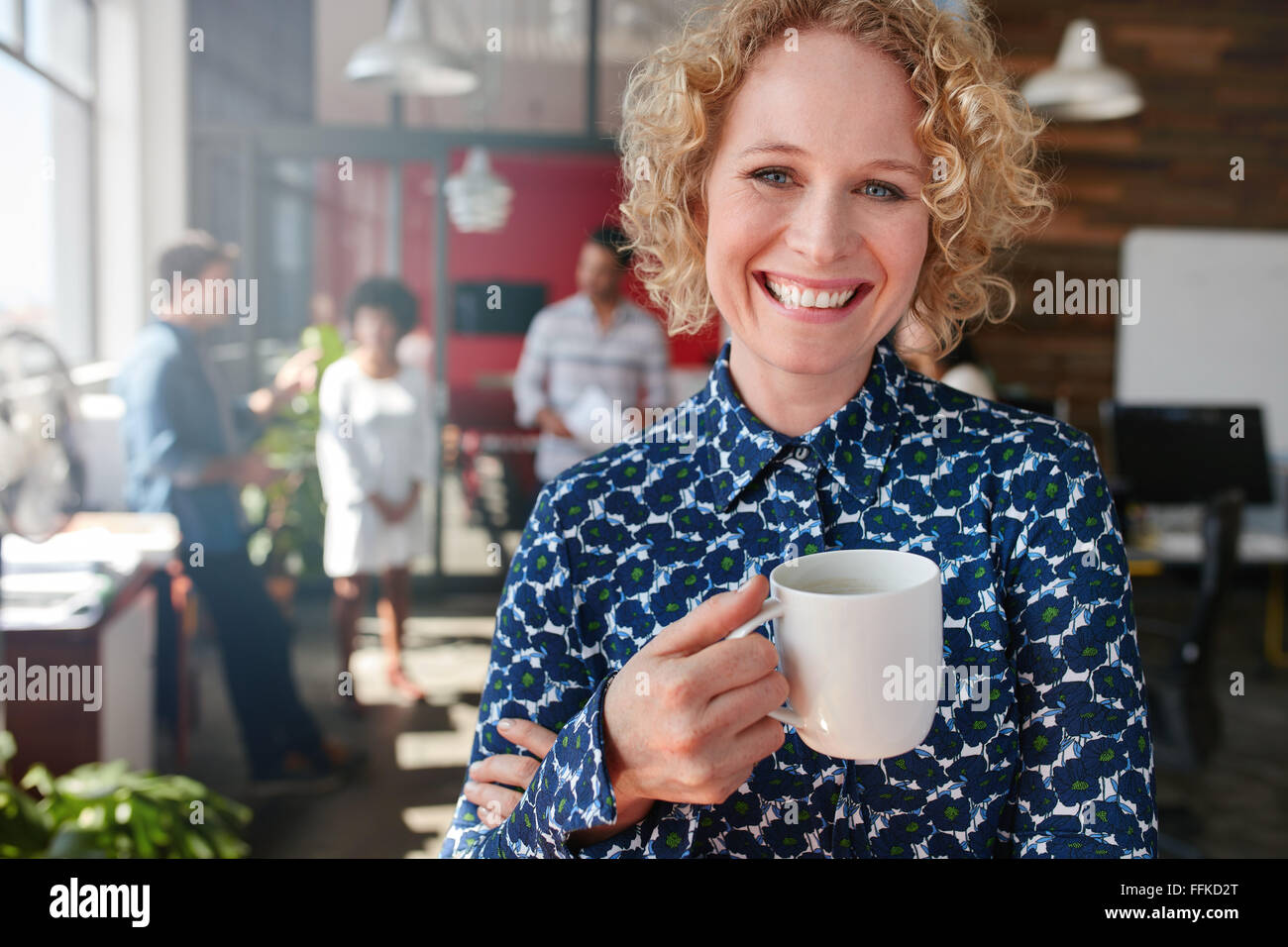 Portrait of cheerful female designer holding a cup of coffee. She is standing in office with colleagues discussing in background Stock Photo