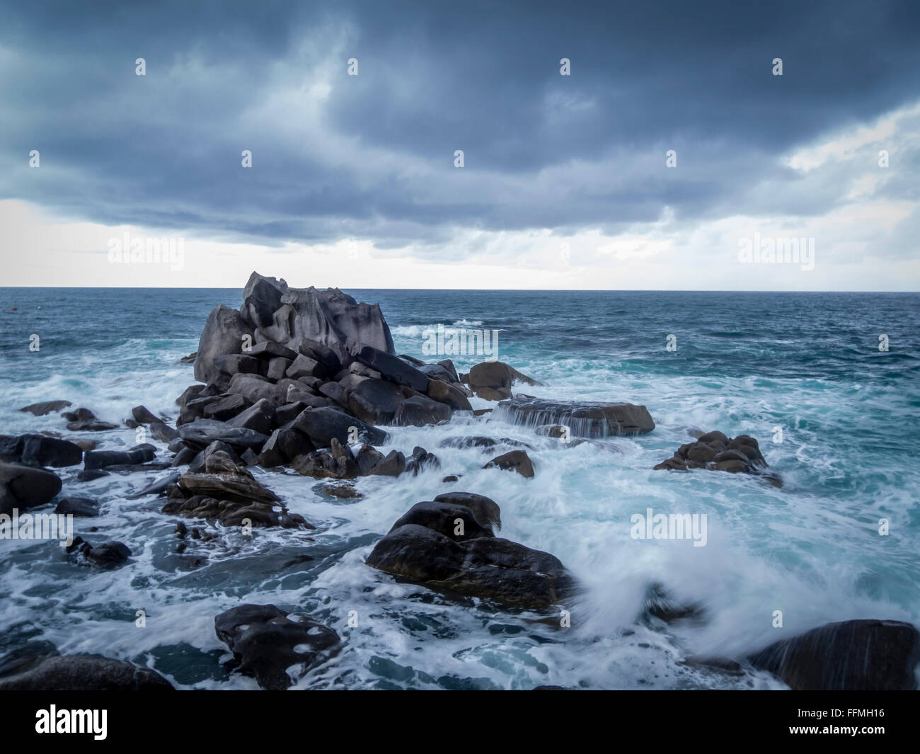Waves crash over rocks with dark clouds looming overhead Stock Photo