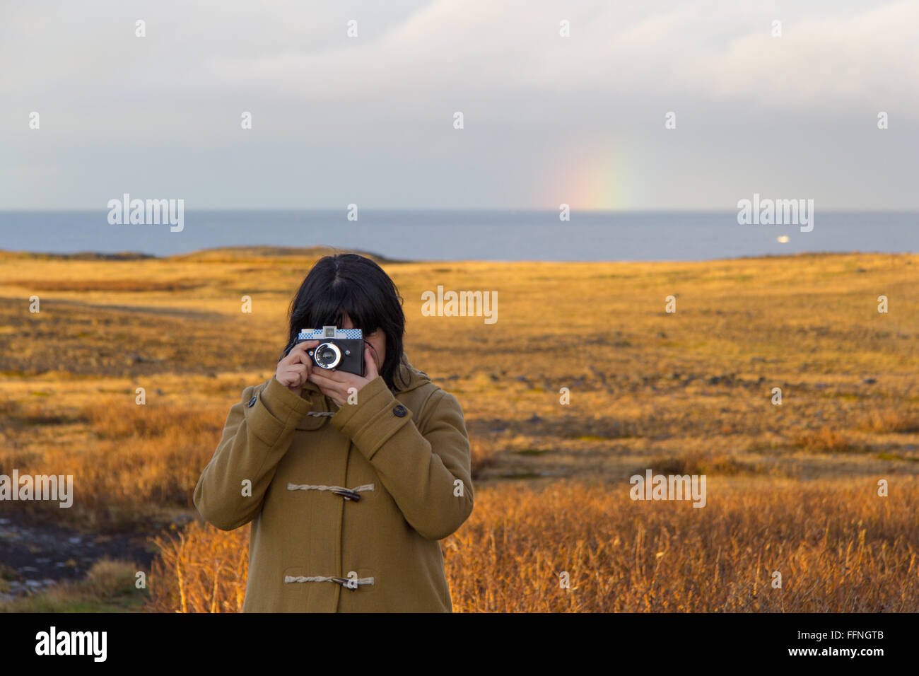 A woman takes a picture in a sunny afternoon in Snaefellsjoekull peninsula, Iceland, with a Diana camera and the rainbow behind Stock Photo
