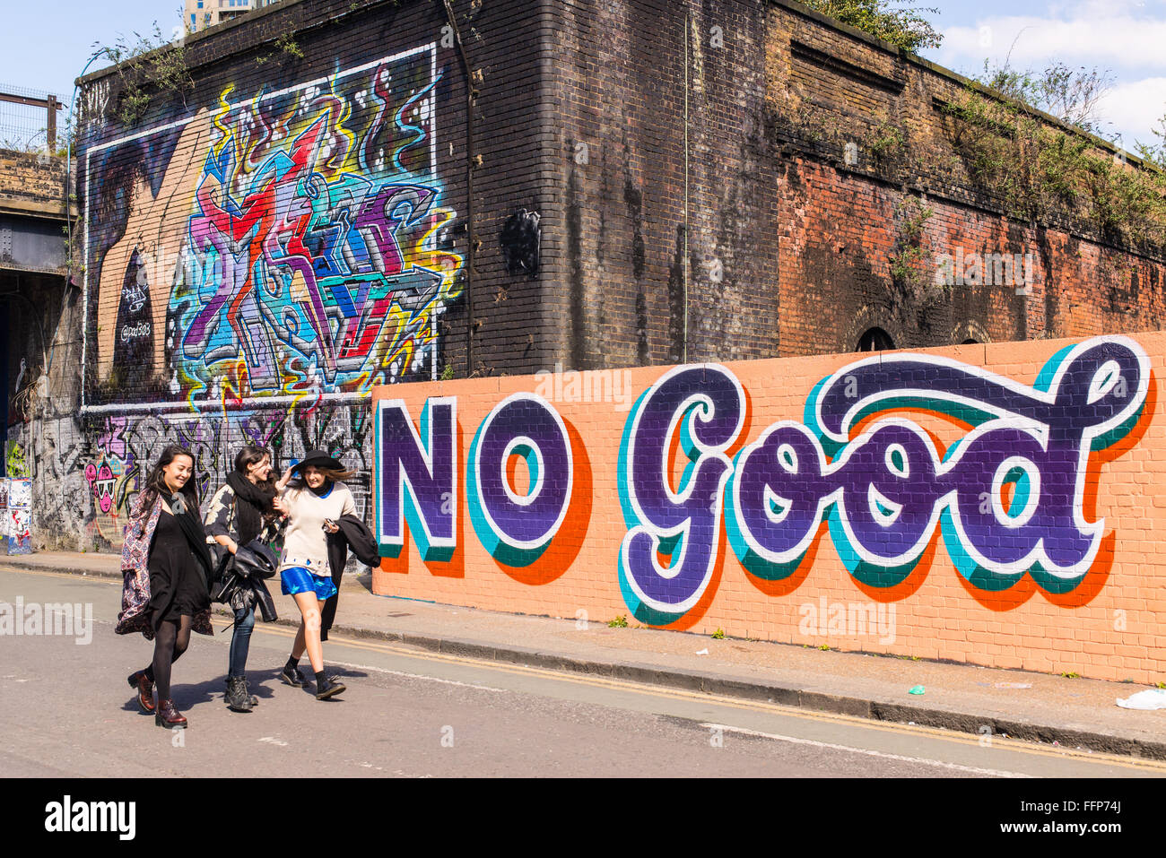 Three hipster girls walking in front of a wall covered in graffiti with the writing 'No Good' Stock Photo