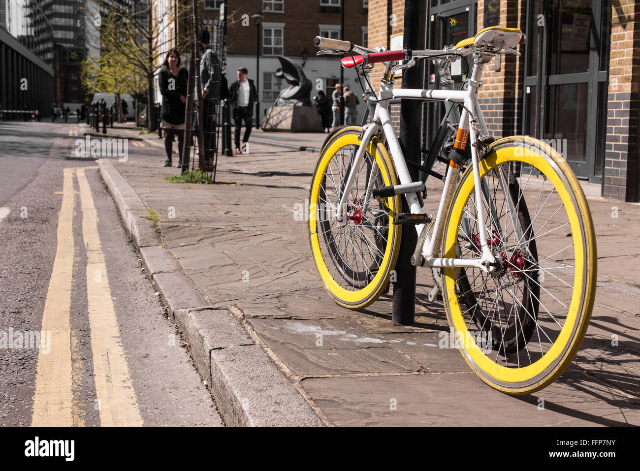 Modern single gear bicycle with yellow tyres locked to a street lamp. Blurred background with people walking in a street. Stock Photo