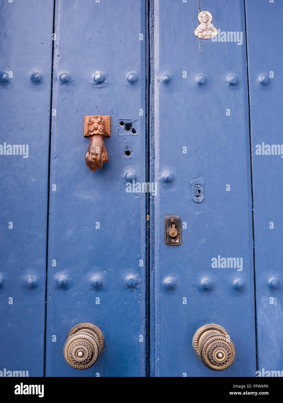Front Door, Blue, panelled, with hand shaped knocker, a pair of door knobs, a religious symbol and a euro lock, Frigiliana, Malaga, Andalusia, Spain. Stock Photo