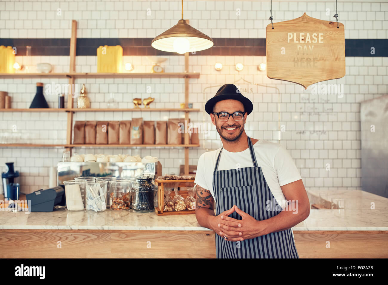 Portrait of happy young man wearing an apron and hat leaning to a cafe counter. Male barista standing at coffee shop looking at Stock Photo