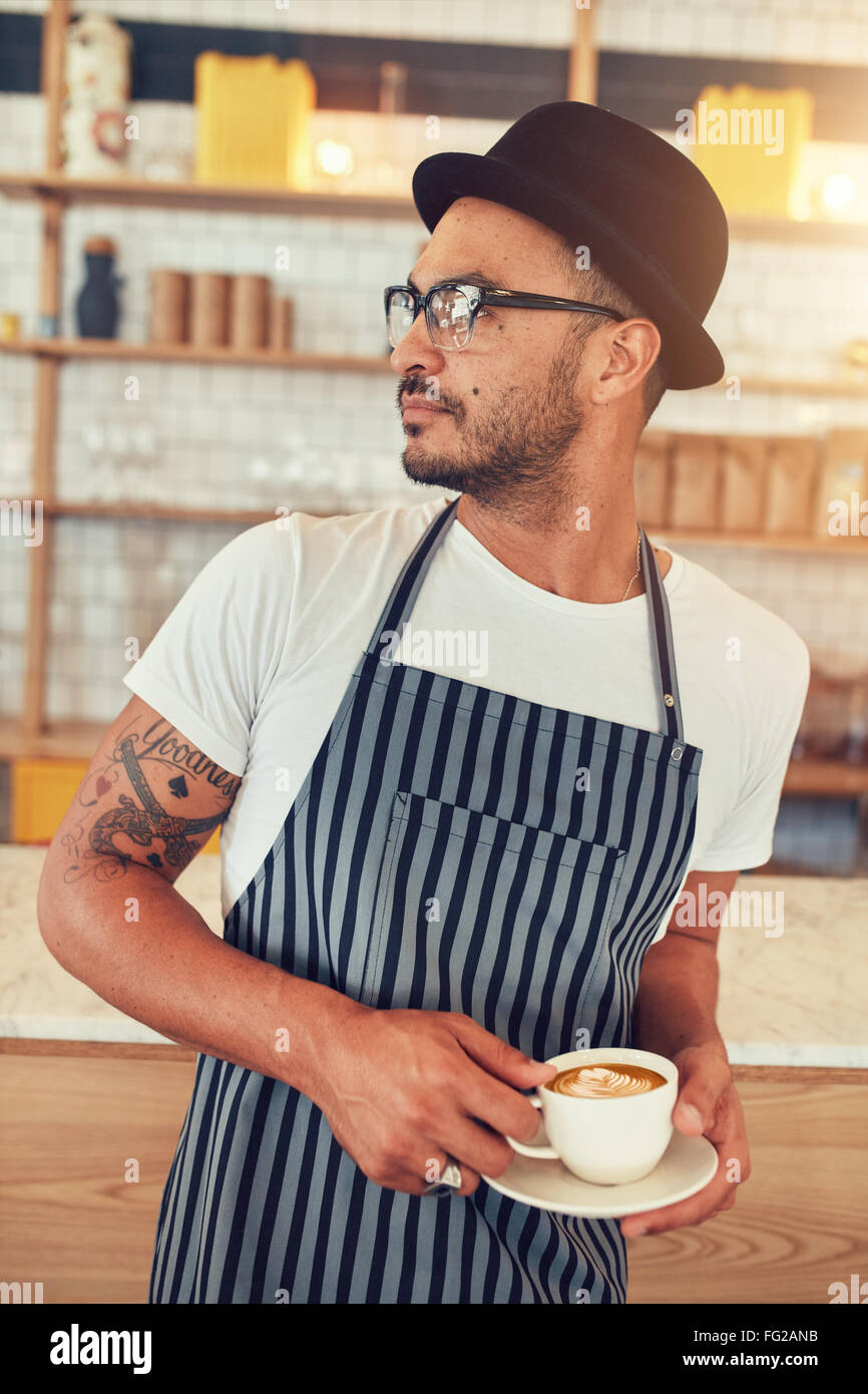 Portrait of young man working in coffee shop standing at bar counter and looking away. Male barista standing at coffee shop with Stock Photo