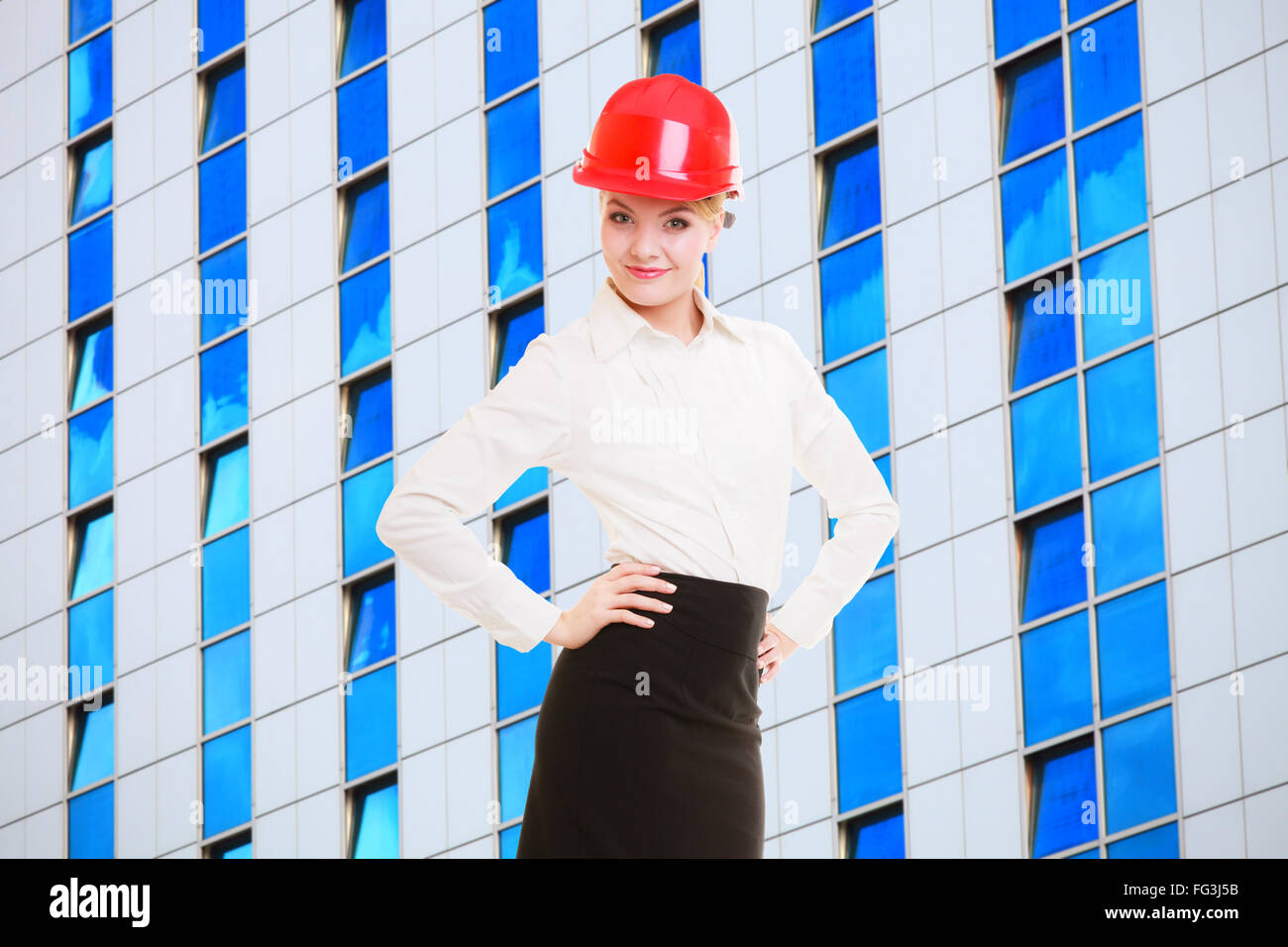 Attractive female engineer and modern futuristic glass building. Woman architect in red safety helmet. Construction site. Stock Photo