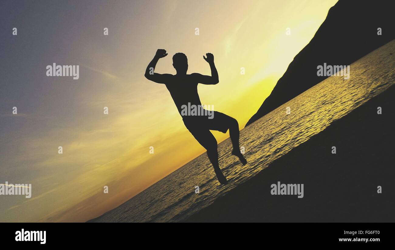 Tilt Image Of Silhouette Man Jumping At Beach Against Sky During Sunset Stock Photo