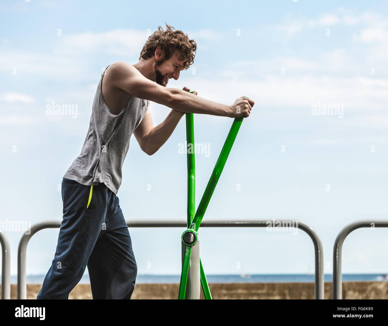 Active young man exercising on elliptical trainer machine. Muscular sporty guy in training suit working out at outdoor gym. Sport fitness and healthy lifestyle concept. Stock Photo
