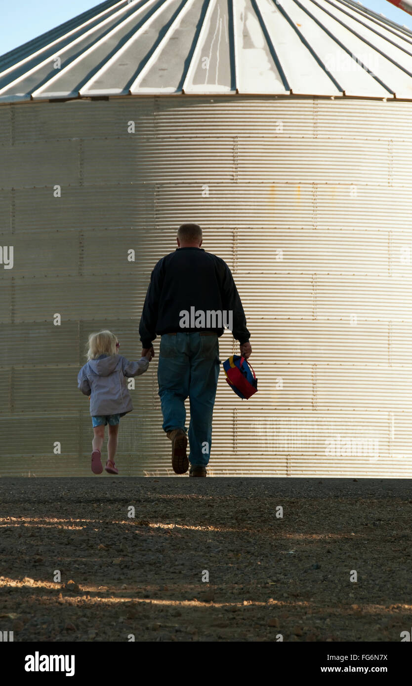 Agriculture - A farmer and his young daughter walk hand-in-hand through his farm yard with a grain bin in the background / near Sioux City, Iowa, USA. Stock Photo