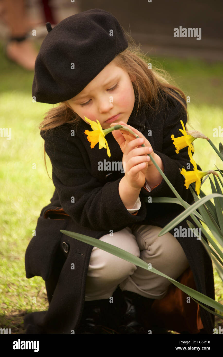 Portrait Of Young Girl Dressed In A Brown Overcoat, Trousers And Beret Is Holding A Yellow Daffodil With One Hand And Stroking It With The Other, C... Stock Photo