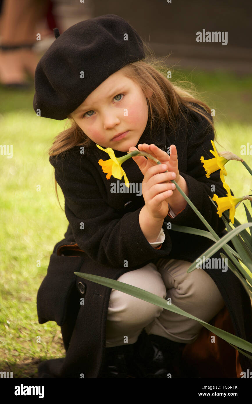 Portrait Of A Young Girl Dressed In A Brown Overcoat, Trousers And Beret Holding A Yellow Daffodil With One Hand And Stroking It With The Other, Cr... Stock Photo